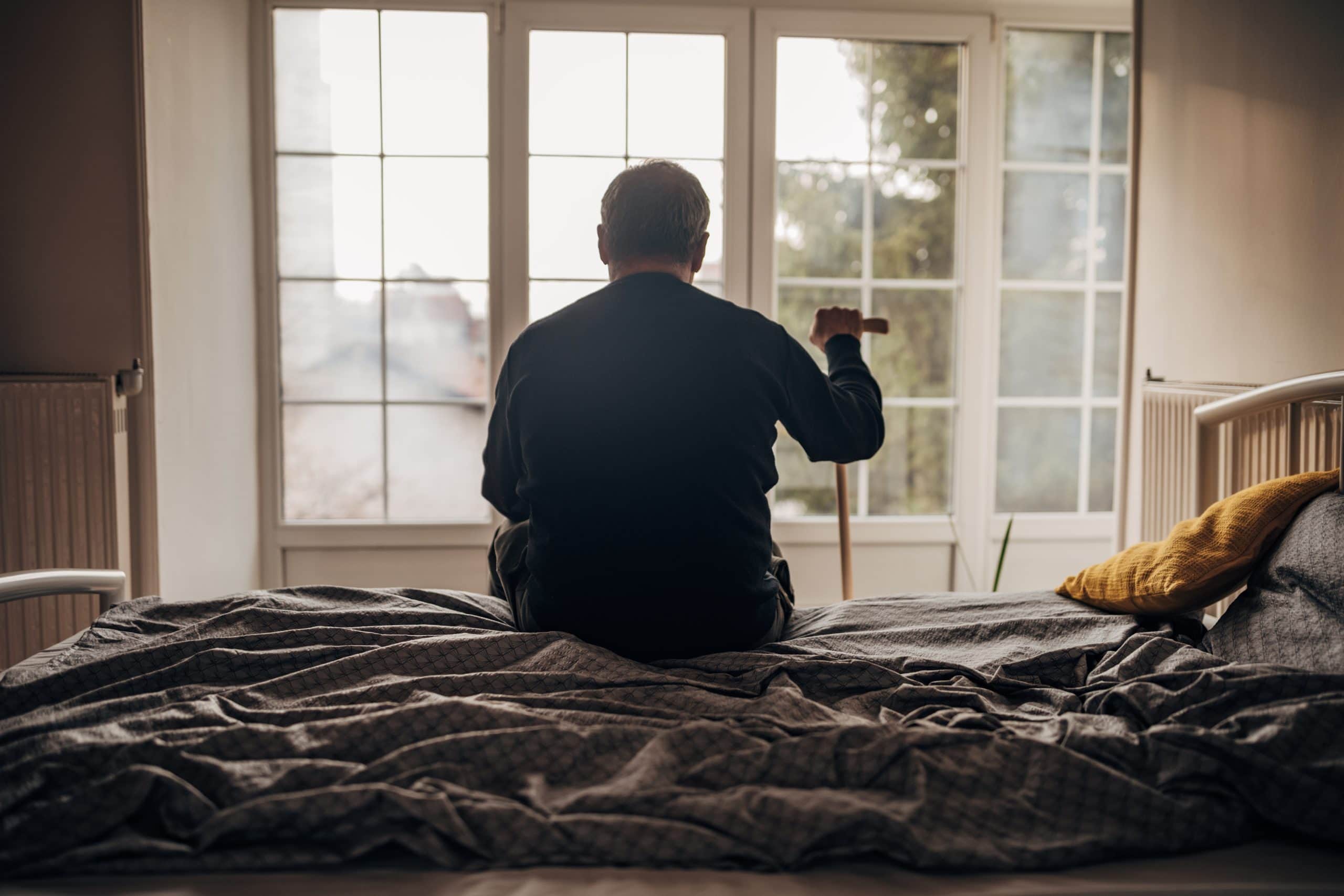 Elderly man sitting on bed with cane, gazing out window in assisted living.