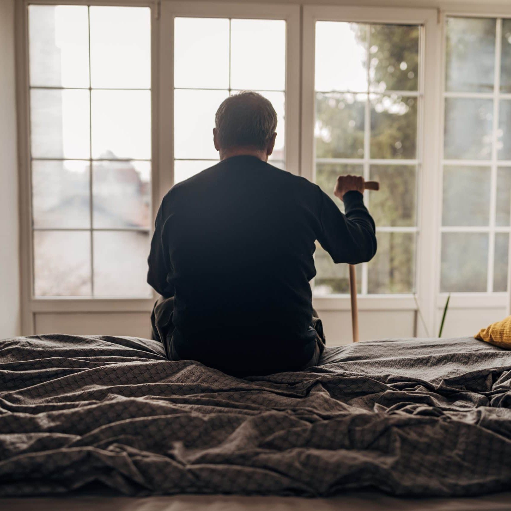 Elderly man sitting on bed with cane, gazing out window in assisted living.